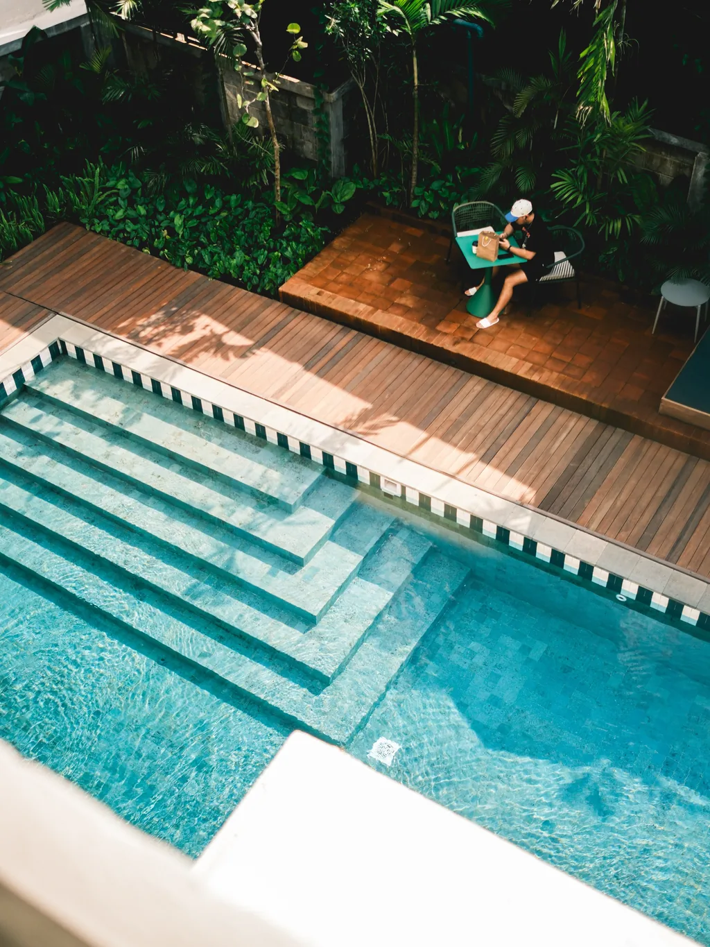Kolam renang bersama dilihat dari atas, dengan tangga berubin, dek kayu, daybed, dan pohon palem