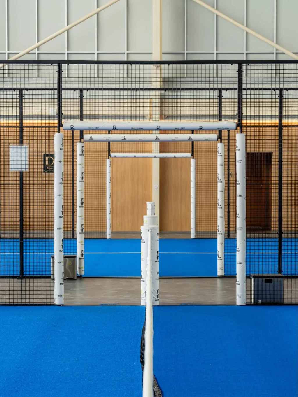 A padel court with blue turf, mesh walls and net posts, seen from one end