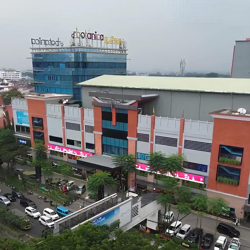 Foto udara D'Botanica Bandung Mall: menara kaca biru dengan tulisan d'botanica di atasnya, dan fasad mal bata merah dengan papan neon merah muda serta pohon palem di depannya