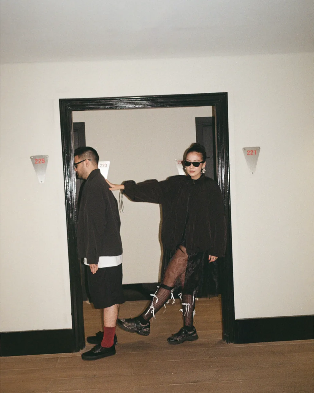 Prewedding couple in dark outfits at an open doorway in a hotel corridor, she leaning on the door frame in sunglasses