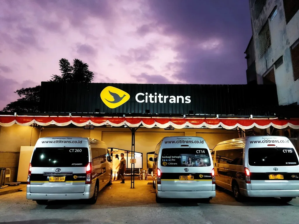Cititrans Pasteur saat senja: tiga mobil shuttle putih terparkir di depan papan nama Cititrans yang menyala