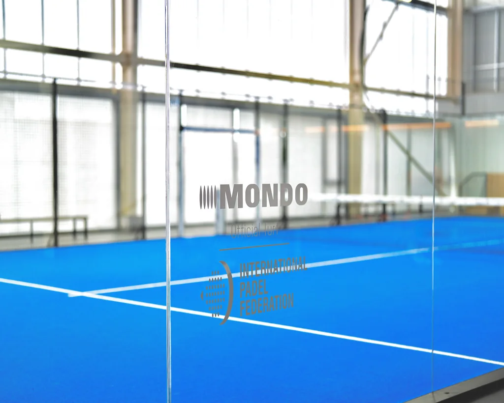 Lapangan padel biru terlihat dari balik dinding kaca bertanda Mondo